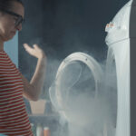 Upset,Woman,Staring,At,Her,Broken,Washing,Machine,,The,Room